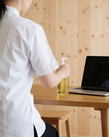A therapist in a white uniform prepares massage oil on a wooden table in a spa setting with wooden walls.