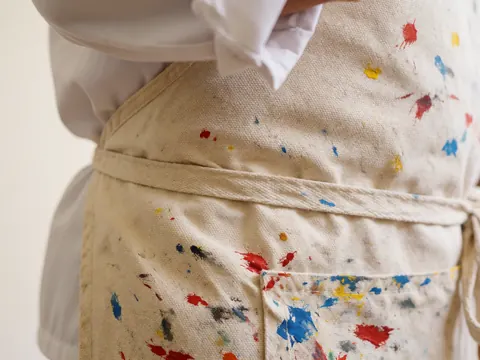 Close-up of a canvas apron belt featuring red, blue, and yellow paint splatters, highlighting artistic tools in a painting studio setting.
