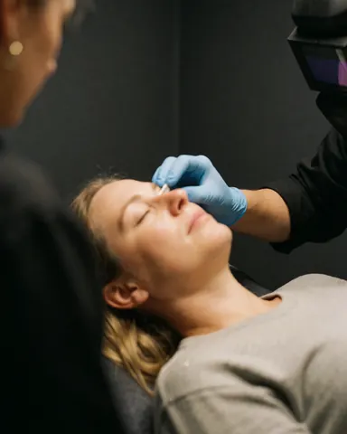 Close-up of a technician wearing blue gloves applying ointment near closed eyes during a microblading aftercare session in a salon with gray walls