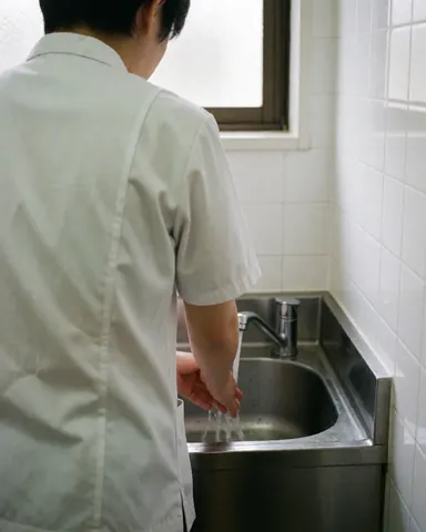Shiatsu professional washing hands in a stainless steel sink with white tiled walls and natural light from a window