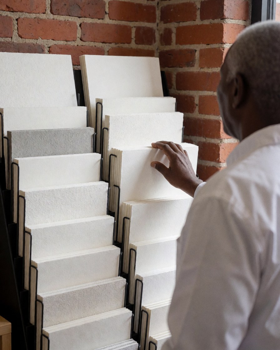 A customer examines various textured papers displayed on a shelf in a stationery store with a red brick wall background.