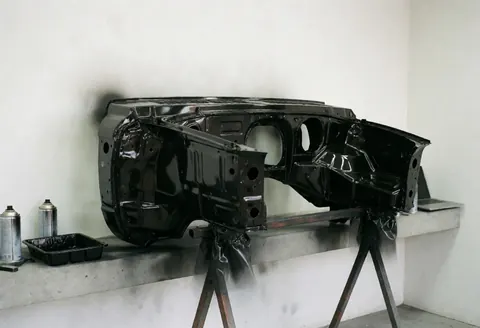 Freshly painted glossy black car body frame resting on a metal stand in a workshop with spray cans and a paint tray nearby