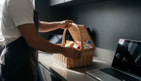 A man opens a wicker welcome basket containing snacks and water in a modern kitchen with gray walls.