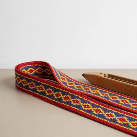 Woven belt with red, yellow, and blue geometric patterns on a beige surface next to a wooden tool for craft workshops