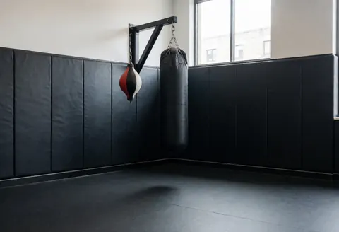 Corner of a boxing gym featuring a heavy bag and speed bag, padded walls, and rubber flooring for professional training.