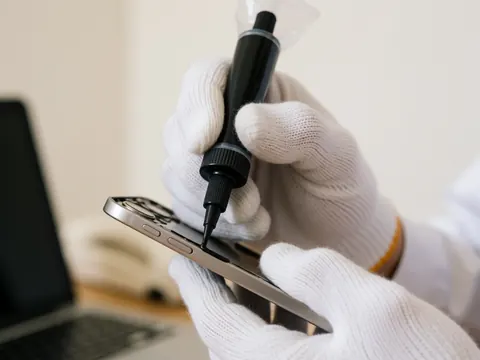 Close-up of a gloved hand applying black adhesive to the edge of a smartphone against a white wall