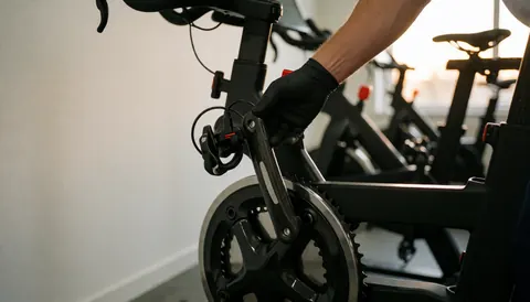 Close-up of a hand adjusting a power meter sensor on a stationary bike in a gym with white walls and sunset light