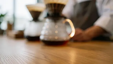 A glass coffee carafe sits on a wooden table with a drip filter nearby, providing a clean background for coffee shop marketing.