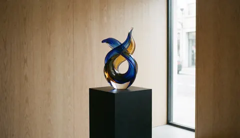 Abstract glass sculpture with blue and yellow ribbons on a black pedestal in a room with wooden walls and natural light