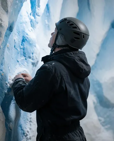 A climber wearing a helmet and dark jacket evaluates a blue and white ice wall during an indoor climbing class.