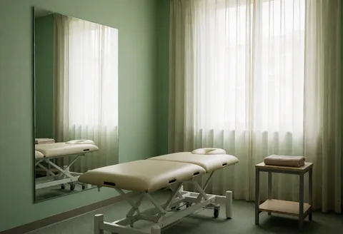 Adjustable treatment bed in a green-walled physiotherapy room with a large mirror, curtains, side table, and towel.