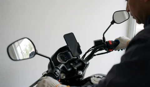 Close-up of a motorcycle taxi handlebar with a smartphone secured in a holder, showing a gloved hand and white wall background