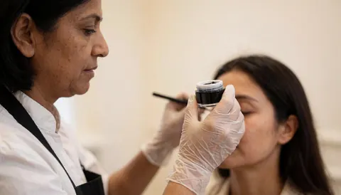 Lash technician wearing gloves applies tint to a client with closed eyes in a salon with cream walls