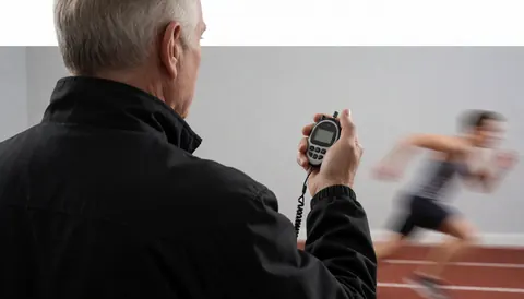 Soccer coach holding a digital stopwatch while watching an athlete run on a track, viewed from behind