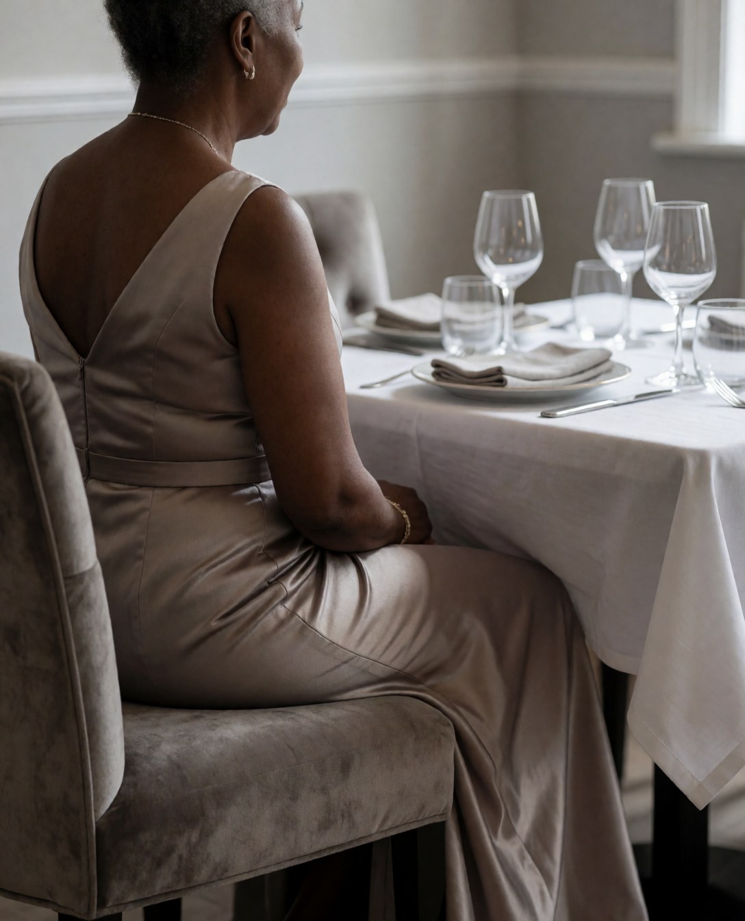 A woman in a formal dress sits on a velvet chair, looking at an elegant table with dinnerware and wine glasses in a light gray restaurant.