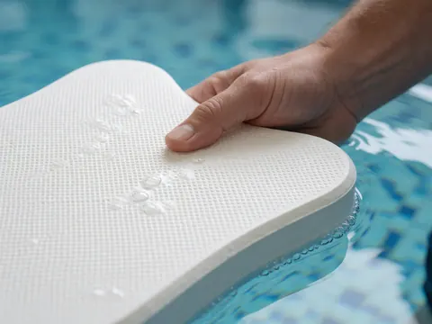 Close-up of an adult hand gripping a textured white swimming kickboard partially submerged in clear blue pool water with bubbles over blue tiles