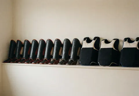 Shin guards and chest protectors neatly arranged on a shelf in a dojo with cream-colored walls.
