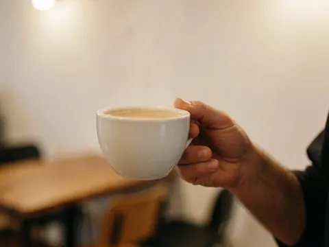 A hand holding a white cup of coffee with rising steam in a warm cafe setting