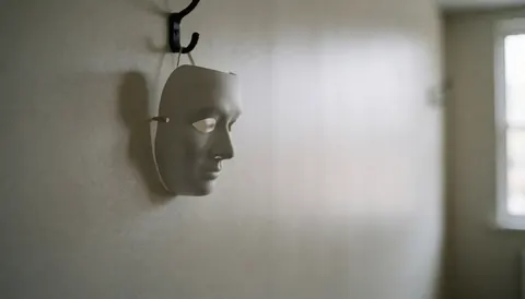 A white theatrical mask suspended on a hook against a textured wall with soft lighting and a blurred background.