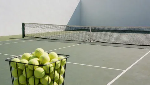 Tennis court featuring a basket of yellow balls, net, and white walls, suitable for tennis instructor business websites