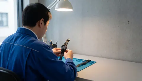 Watchmaker adjusting a digital watch on a blue mesh surface in a workshop with concrete walls and text space