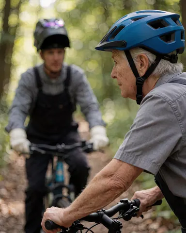 A senior man riding a mountain bike with a blue helmet, followed by his coach on a forest trail.