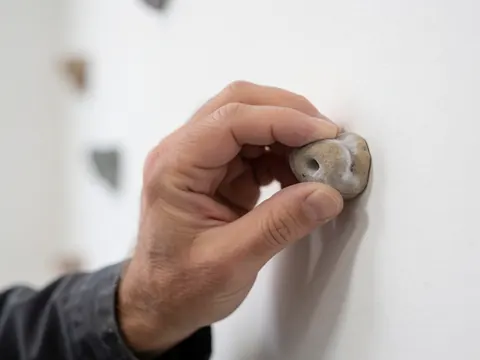 Close-up detail of an adult hand gripping a climbing hold on a white wall, highlighting the texture and action of indoor climbing.