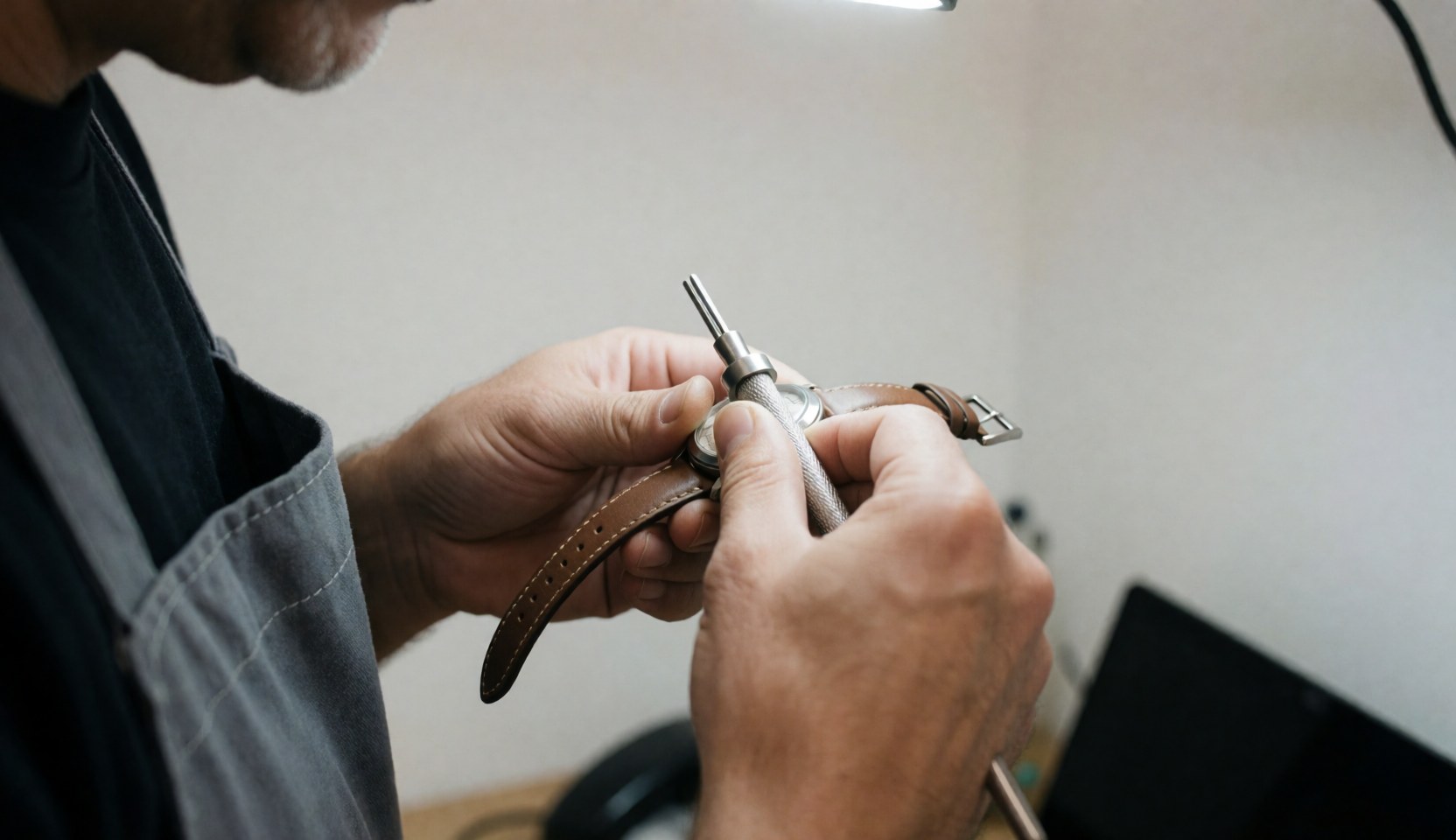 Close-up of hands using a metal tool to repair a watch with a leather strap in a workshop with a white wall.