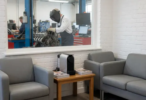 Waiting area with gray chairs and coffee machine, mechanic working on car engine through glass window