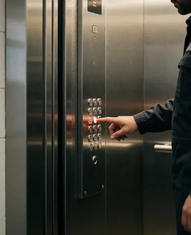 Technician in uniform pressing control button inside elevator with stainless steel walls and warm lighting, showing elevator repair service