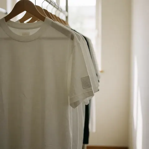 White t-shirt with embroidered patch on sleeve hanging on wooden hangers in a clean dressing room with white walls and natural light