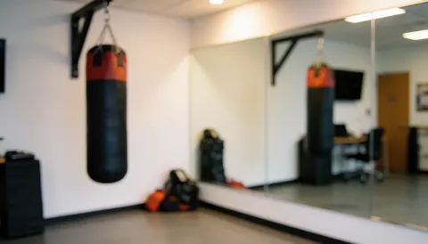 Red and black punching bag hanging in a boxing studio with white walls, large mirror, gray floor, and warm lighting for text background.