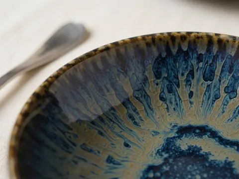 Close-up of a handmade ceramic bowl with blue and gold crackled glaze resting on cream-colored tablecloths