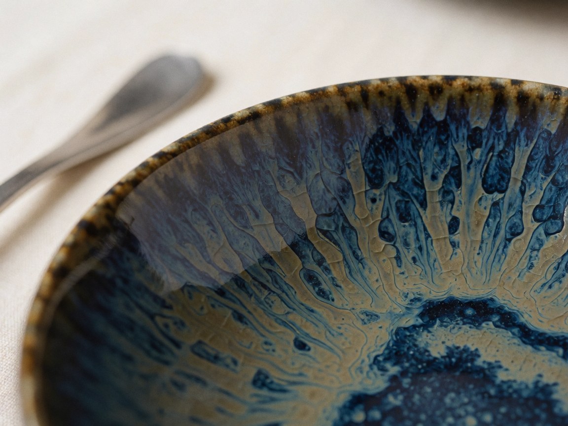 Close-up of a handmade ceramic bowl with blue and gold crackled glaze resting on cream-colored tablecloths