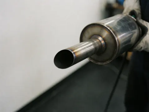 Detailed view of an automotive exhaust muffler showing weld seams, held by gloved hands in a workshop setting.