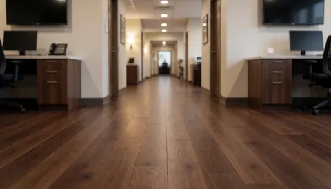 Commercial hallway featuring dark hardwood flooring, warm lighting, and office furniture against white walls, suitable as a background for text.