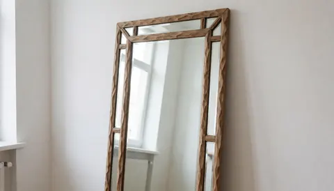 A handmade mirror featuring a carved wooden frame, reflecting natural light and a white wall in a glassblowing workshop setting.