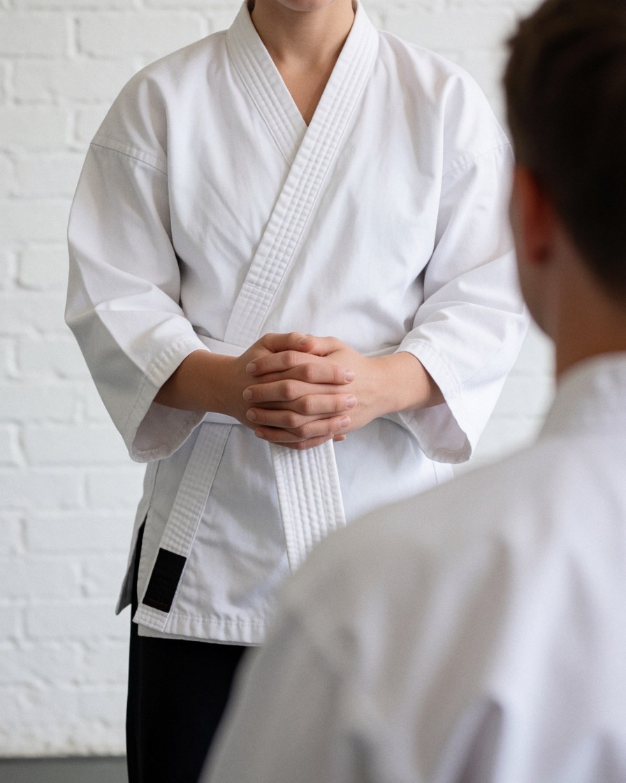 Karate instructor hands a white belt to a student in a dojo with a white brick wall, showing respect with clasped hands