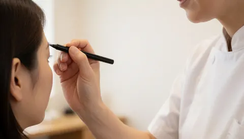Technician in white uniform applying black pencil to client's eyebrow against white background