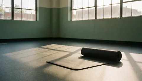 A rolled black yoga mat sits on a textured rubber gym floor, illuminated by morning light from industrial windows in an empty fitness space.