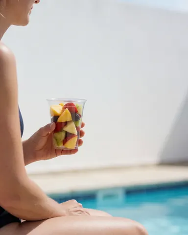 A woman relaxes by the poolside enjoying fresh fruit under bright sunlight with blue water and white walls.