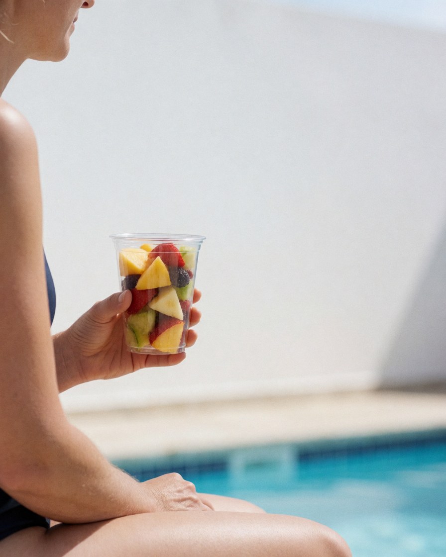 A woman relaxes by the poolside enjoying fresh fruit under bright sunlight with blue water and white walls.