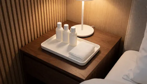 White tray with bath products and towel on wooden nightstand beside bed with white sheets and slatted wall