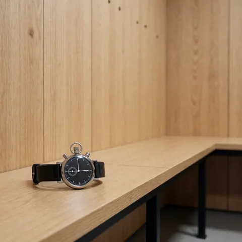 Black stopwatch with leather strap resting on a wooden bench in a modern locker room with light wood paneling