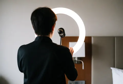 A makeup artist adjusts a ring light in a hotel room with neutral walls and a bed headboard, preparing equipment for an event.