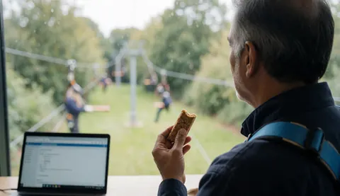 A man wearing a blue harness looks at a zipline while holding an energy bar, with a laptop on the table nearby.