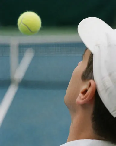 Tennis player wearing white cap watching ball in air, tennis net visible, blue court background, ideal for training services