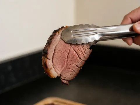 A piece of grilled meat held by metal tongs against a dark kitchen background with warm white walls and a glossy fat texture.