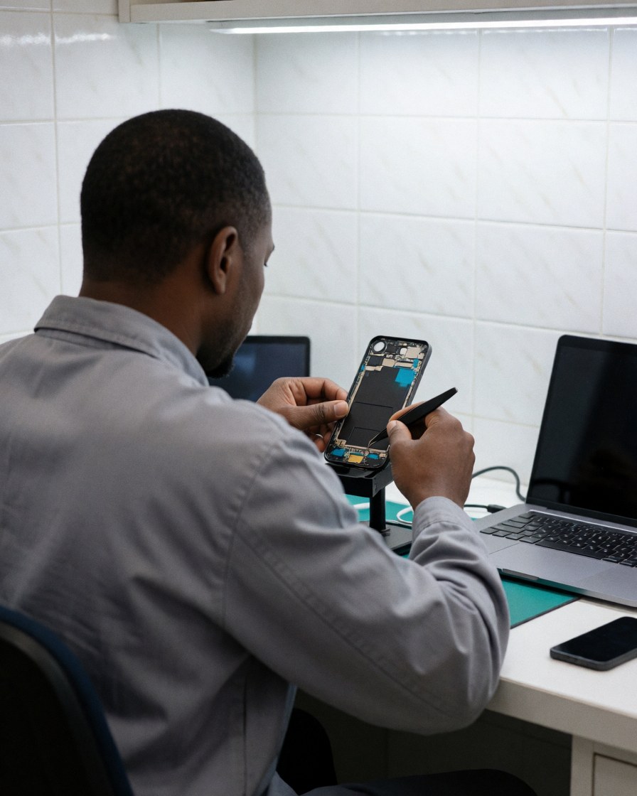 Technician using tweezers to repair a disassembled smartphone in a workshop with white tiled walls and a laptop nearby