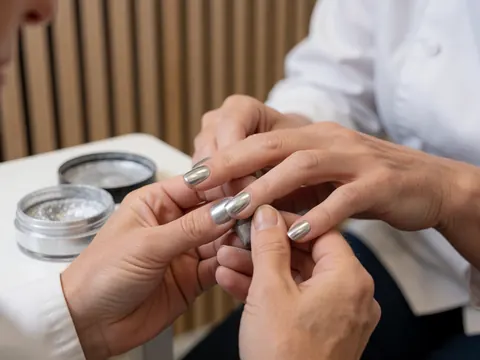 Professional manicure applying metallic powder to nails with tools and light wood walls in the background